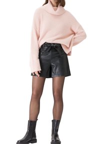 Helles rosa, fuzziges Sweater mit einem Cowl-Neck, kombiniert mit hoch taillierten, schwarzen Leder-Shorts, die an der Taille gebunden sind. Schwarze Knöchelstiefel runden das Outfit ab.