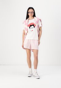 Hvit T-skjorte med Frida Kahlo-grafikk, rosa-hvite stripede cardigan, stripete shorts, hvite sneakers med snøring og briller.