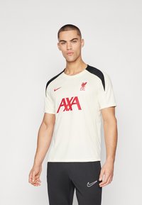 Uomo che indossa una maglietta bianca del Liverpool FC con il logo rosso AXA e accenti neri sulle spalle, abbinata a pantaloni neri Nike.