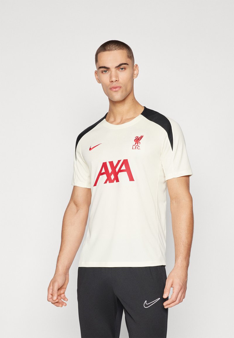 Uomo che indossa una maglietta bianca del Liverpool FC con il logo rosso AXA e accenti neri sulle spalle, abbinata a pantaloni neri Nike.