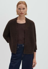 Donna con capelli corti biondi che indossa un cardigan aperto di colore marrone scuro e dalla trama accentuata sopra una maglia scura e jeans blu scuro a vita alta, su uno sfondo semplice.