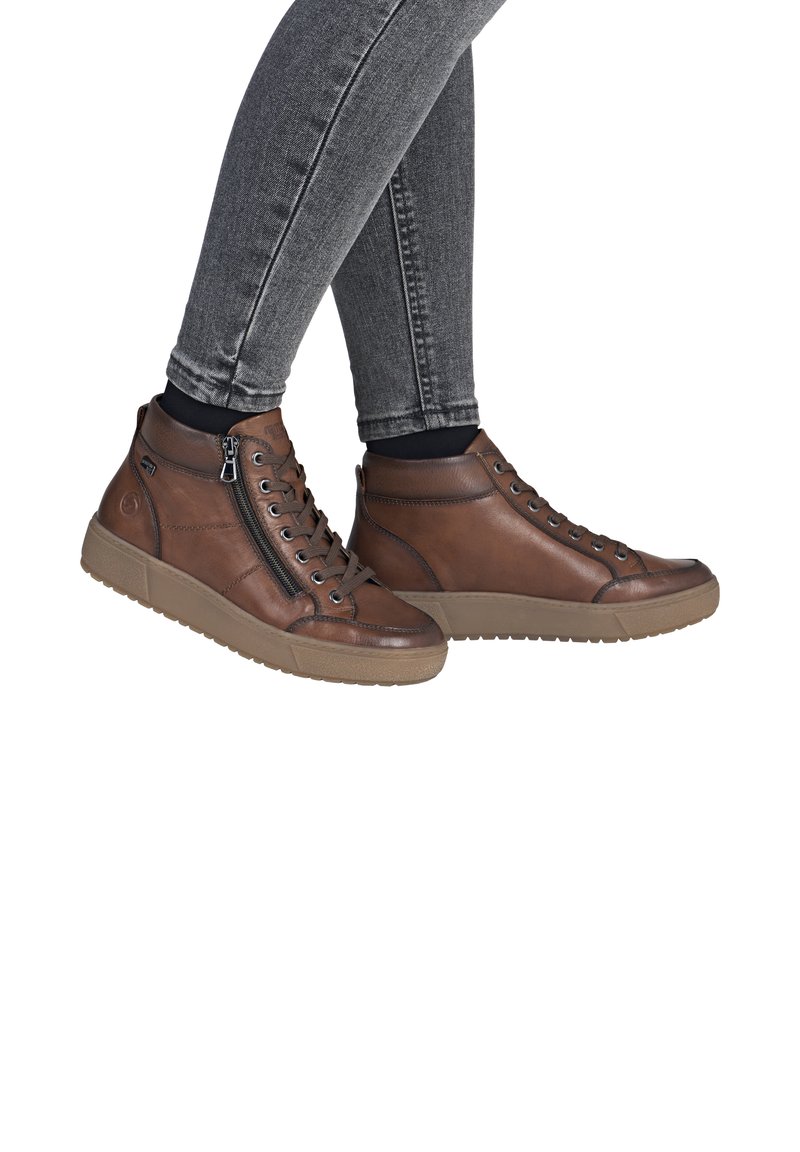 Baskets montantes en cuir brun avec une surface texturée, fermeture éclair latérale et semelle en caoutchouc, associées à un jean slim gris foncé.