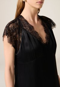 Zwarte zijden jurk met een diepe V-hals en kanten mouwkapjes. Het kant heeft bloemmotieven en een verfijnde afwerking langs de randen.