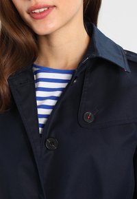 Marinblå trenchcoat i glansigt tyg med klassisk krage. Har två svarta knappar med röda detaljer och en randig skjorta under.