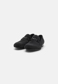 Chaussures noires à enfiler avec une finition texturée, bout rond, petit talon, et détail de bande décorative sur le dessus. Surface lisse.