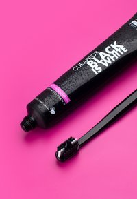 Curaprox Black is White tandkrämstub, svart med grå mönster, bredvid en svart tandborste med en blandning av vita och svarta borst, på rosa bakgrund.