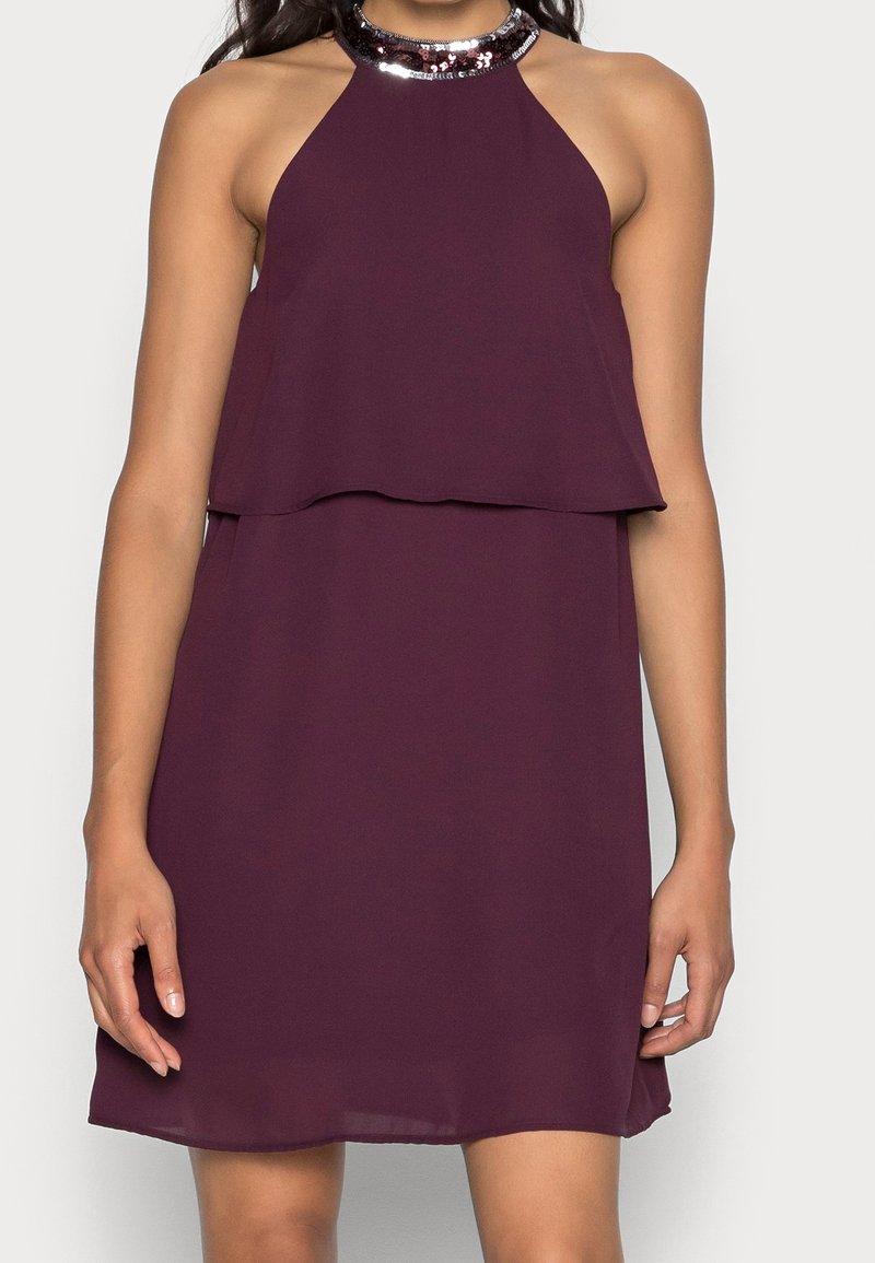 Femme portant une robe parée sans manches couleur prune, avec un col halter orné de sequins, un corsage à couches et un ourlet atteignant le genou, sur un fond clair.