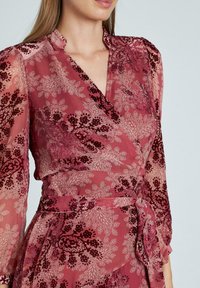 Rosenrotes Wickelkleid aus transparenter, gemusterter Stoff. Mit floralen Designs, langen Ärmeln, einem gebundenen Bund und gerüschten Akzenten an den Manschetten.