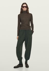 Vrouw in een bruine coltrui, losse donkergroene joggingbroeken, zwarte instappers en grote bruine zonnebril, staand tegen een effen achtergrond.