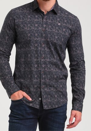 Man wearing dark patterned long-sleeve button-up shirt and dark jeans, left hand partially in pocket, against plain background.