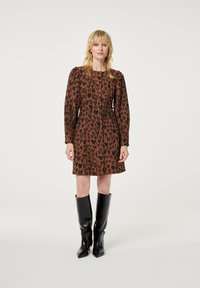 Braunes Kleid mit schwarzem Herzmuster, langen Ärmeln und tailliertem Oberteil; kombiniert mit glänzenden schwarzen Stiefeln bis zum Knie mit spitzen Zehen.