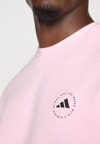 Chemise rose clair à manches courtes avec un logo noir rond portant le texte "ADIDAS GOLF" et un design à trois bandes sur le devant. Texture lisse.