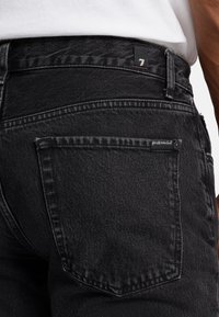 Des jeans en denim noir présentent une poche arrière avec un bord cousu et une étiquette. Le tissu a une texture lisse et un motif de décoloration subtil.