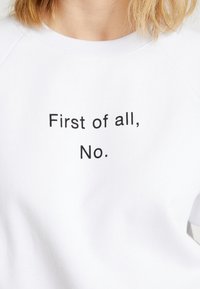 Sweatshirt blanc avec le texte "D'abord, non." en lettres noires en gras. Le matériau est doux avec une texture lisse et un col côtelé.