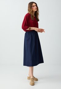 Femme debout portant un chemisier rouge à manches longues, une jupe midi bleu marine, des baskets beiges et un bracelet épais à son poignet droit.