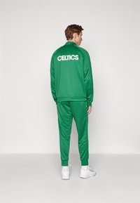 Zeleni trenirku koji se sastoji od jakne na patentni zatvarač i trenerki. Jakna ima natpis "CELTICS" u bijeloj boji na leđima. Glatka tkanina, elastične manžete.