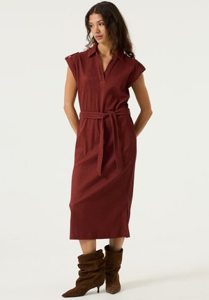 Vrouw met een mouwloze, gekraagde, met een riem getailleerde bordeauxrode midi-jurk en slouchy bruine laarzen, staand en opzij kijkend.
