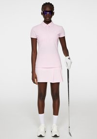 Różowa koszulka polo do golfa z kołnierzykiem, w zestawie z pasującą spódniczką. Biała rękawica golfowa, czarno-białe buty sportowe i okulary przeciwsłoneczne.