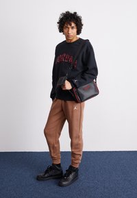 Felpa nera con scritta "JORDAN" rossa, pantaloni della tuta marroni e sneaker alte nere, mentre tiene una piccola borsa nera con dettagli rossi.