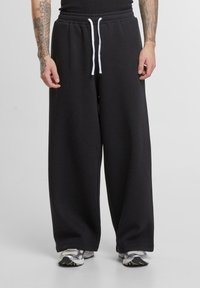 Sorte brede sweatpants lavet af blødt stof, med elastisk talje og snor samt hvidt pynt. Båret med sportssko.