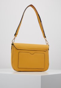 Žlutá kožená kabelka s klopou, nastavitelným popruhem a přední kapsou. Texturovaný povrch a šití podél okrajů.