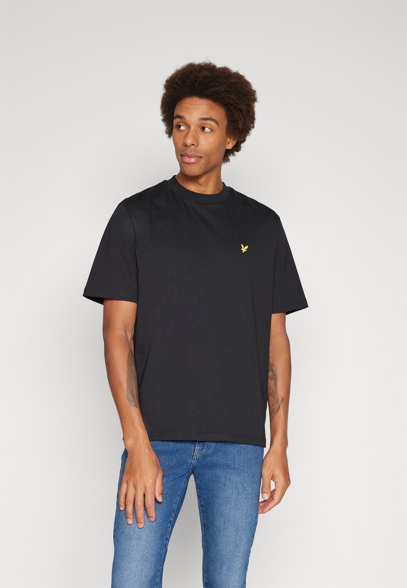 T-shirt nera in cotone a maniche corte e colletto rotondo. Presenta un piccolo logo giallo sul petto. Indossato con jeans blu.