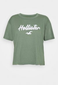 Olivgrön cropped t-shirt i mjukt tyg med en vit broderad "Hollister"-logotyp över bröstet. Korta ärmar och rundad halsringning.