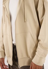 Lys beige zip-up hettegenser laget av mykt materiale, med to frontlommer, ribbede mansjetter og "URTRN" brodert på Ermet.