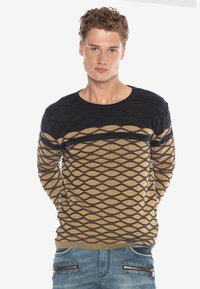 Braun und schwarz gemusterter Pullover mit einem strukturierten Rautendesign, Rundhalsausschnitt und geripptem Saum, kombiniert mit blauen Jeans mit Reißverschlusstaschen.