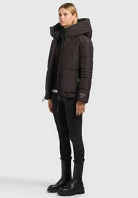 Doudoune noire à col montant et capuche, dotée de poches zippées et d'un design matelassé. Associée à des leggings noirs et des bottes.