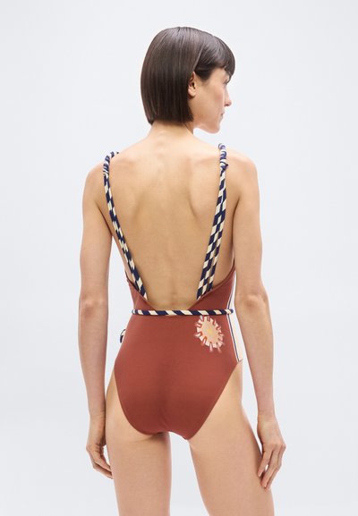 Γυναίκα με κοντά σκούρα μαλλιά, που φοράει ολόσωμο μαγιό σε χρώμα σκουριάς με βαθύ άνοιγμα στην πλάτη, ριγέ λουράκια και μικρό λουλουδάτο σχέδιο στο πλάι.