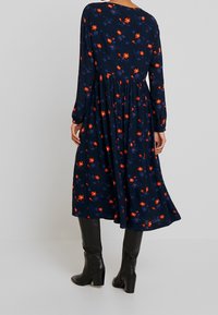 Granatowa sukienka z długimi rękawami, ozdobiona kwiatowym wzorem w odcieniach pomarańczowym i niebieskim, z marszczoną talią i zwiewnym dołem, noszona z czarnymi kozakami za kolano.