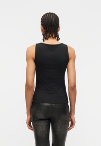 Personne avec des tresses cornrow portant un haut noir sans manches et un pantalon noir texturé, vue de dos sur un fond uni.