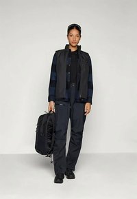 Colete preto por cima de uma camisa xadrez escura, combinado com calças escuras. A pessoa segura uma mochila preta com um design elegante. Fundo neutro.