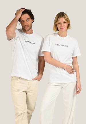 Un homme et une femme debout côte à côte, portant des T-shirts blancs avec le texte « GERTRUDE » et des pantalons de couleur claire sur un fond uni.