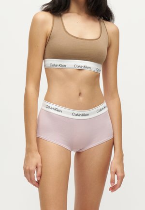 Kvinde iført tan Calvin Klein sports-bh og lyserøde Calvin Klein højtrukne undertøj med hvide elastiske bånd med mærke.