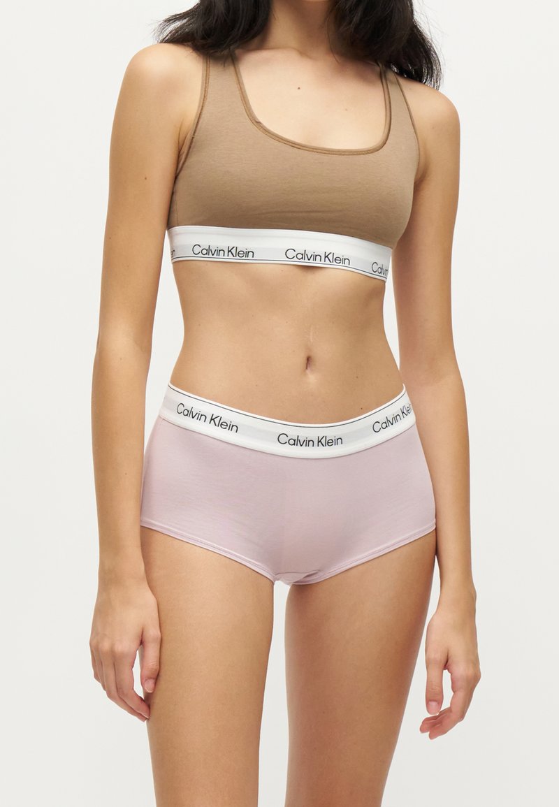 Γυναίκα που φοράει καφέ αθλητικό σουτιέν Calvin Klein και ανοιχτό ροζ ψηλόμεσο εσώρουχο Calvin Klein με λευκές ελαστικές ταινίες με λογότυπο.