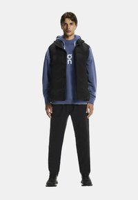 Chaleco acolchado sin mangas negro sobre una sudadera azul con logo, combinado con pantalones negros y zapatos negros. Texturas suaves y colores sólidos.