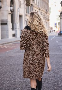 Manteau à imprimé léopard avec un col en fourrure, mi-long, à manches longues, aux couleurs beige et marron, texture douce, porté avec des bottes noires montantes.