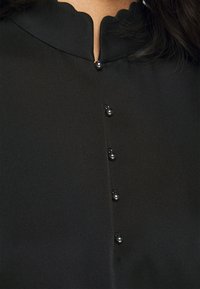 Gros plan sur un chemisier noir à encolure festonnée et cinq petits boutons ronds noirs sur le devant, porté par une personne aux cheveux foncés.
