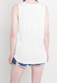 Biała bezrękawnikowa bluzka z okrągłym dekoltem, wykonaną z miękkiego materiału i zaokrąglonym dołem. Połączona z postrzępionymi szortami dżinsowymi.