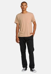 Beige short-sleeve t-shirt with a chest pocket, paired with black pants and black sneakers with white soles. Plain design, casual fit.