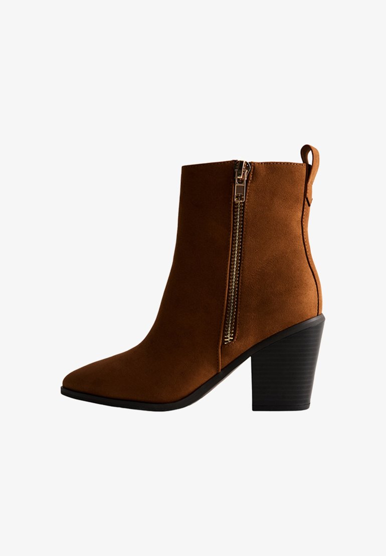 Brązowy zamszowy but za kostkę z ostrym noskiem, bocznym zamkiem błyskawicznym i fakturowanym czarnym obcasem. Czyste linie i smukła sylwetka definiują ten projekt.
