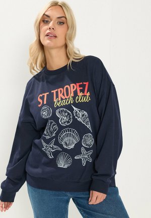 Blonde Frau trägt einen marineblauen Pullover mit dem Schriftzug "ST TROPEZ beach club" und weißen Illustrationen von Muscheln und Seesternen, kombiniert mit blauen Jeans.