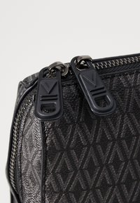 Crna torba teksture s geometrijskim uzorkom, opremljena s dva crna patentna zatvarača s jedinstvenim dizajnom i metalnim dijelom zatvarača.