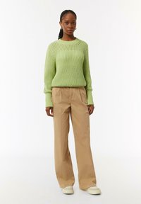Lichtgroene gebreide trui met een textuurpatroon, gecombineerd met beige wijde broek en witte sneakers, staand tegen een effen achtergrond.