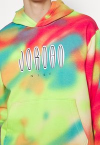 Personne portant un sweat à capuche tie-dye multicolore avec le texte « JORDAN NIKE » sur la poitrine et une poche avant kangourou.