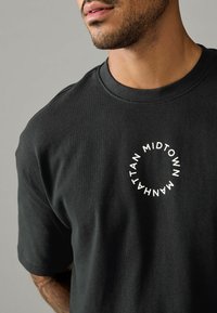 Sort bomulds T-shirt med rund hals, der har en cirkulær hvid grafisk tekst "MIDTOWN MANHATTAN" på brystet. Glat struktur.