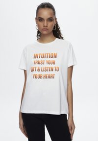 T-shirt en coton blanc avec texte orange : "INTUITION FAIS CONFIANCE À TON INSTINCT & ÉCOUTE TON CŒUR." Manches courtes, coupe décontractée, encolure ronde.