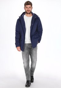 Chaqueta de invierno azul marino con capucha, cremallera frontal, dos bolsillos delanteros y puños elásticos, llevada sobre un suéter gris claro y jeans grises.